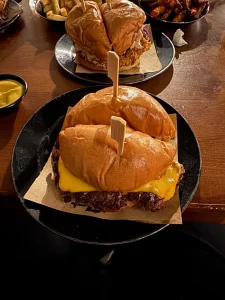 Two cheeseburgers on a black plate with melted cheddar and wooden skewers, fries and dipping mustard nearby on a wooden table.